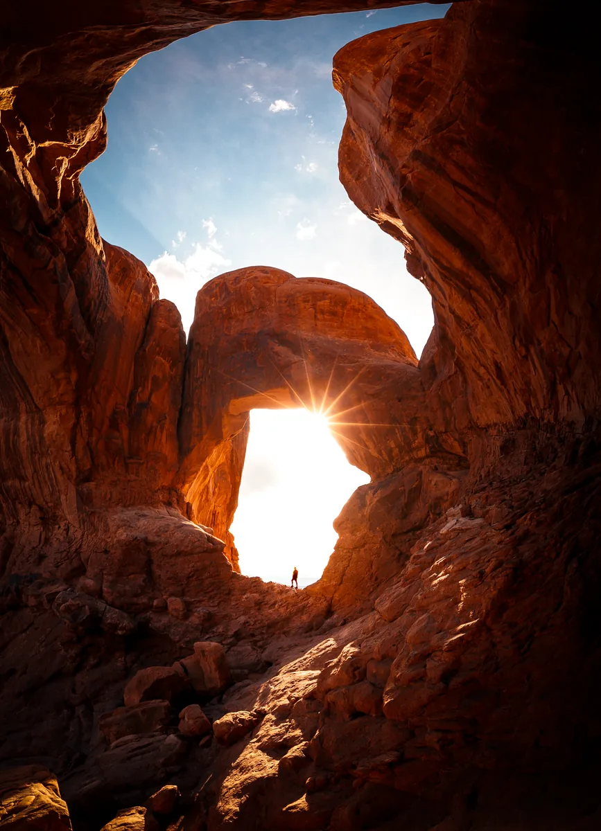 The sun bursting through Double Arch in Arches National Park, Utah, with a tiny figure standing beneath it