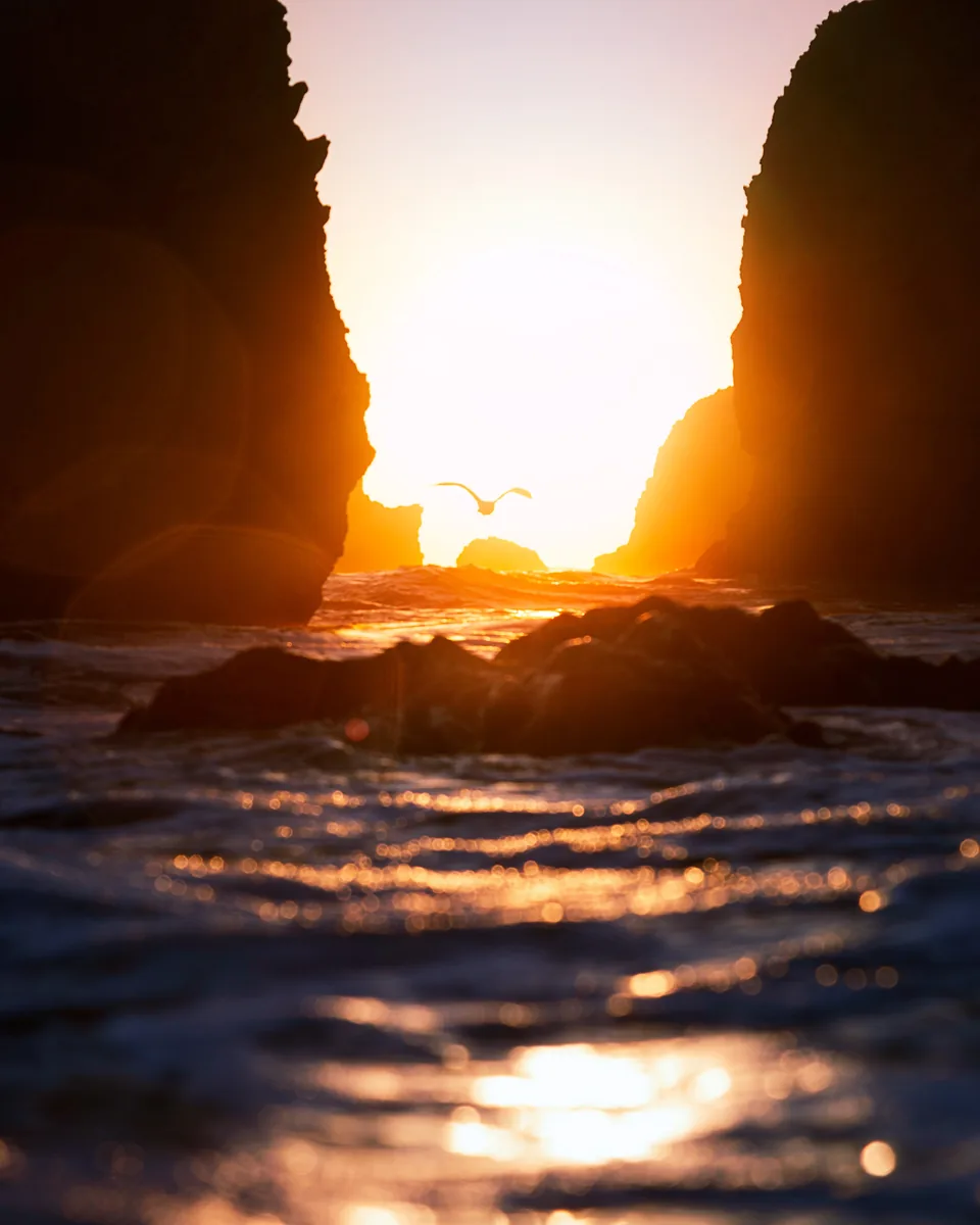The setting sun glowing through a sea arch at El Matador Beach, California, with waves washing over the rocks