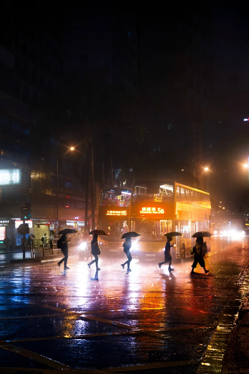 People with umbrellas crossing a rain-soaked street at night in Hong Kong, reflections glowing on the pavement