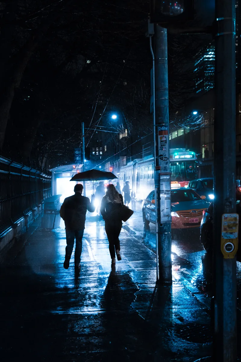 Two people walking under umbrellas through blue light on Queen Street, Toronto, at night