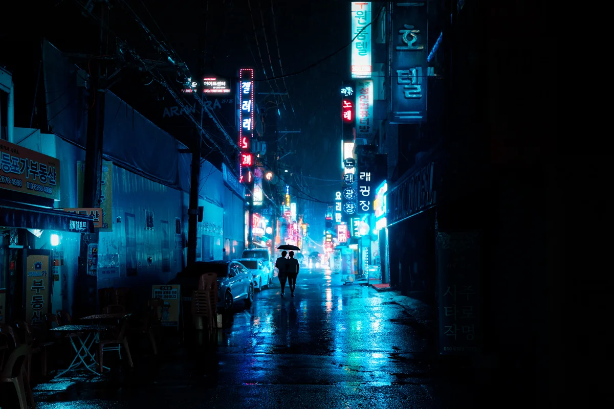 A lone figure with an umbrella walking down a blue-lit Seoul alley on a rainy night