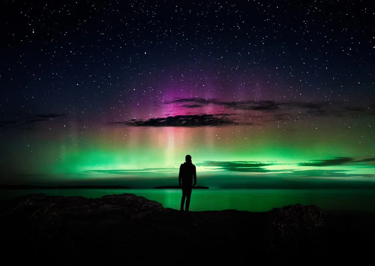 Found, a figure standing beneath green and pink northern lights on the shore of Georgian Bay in Tobermory