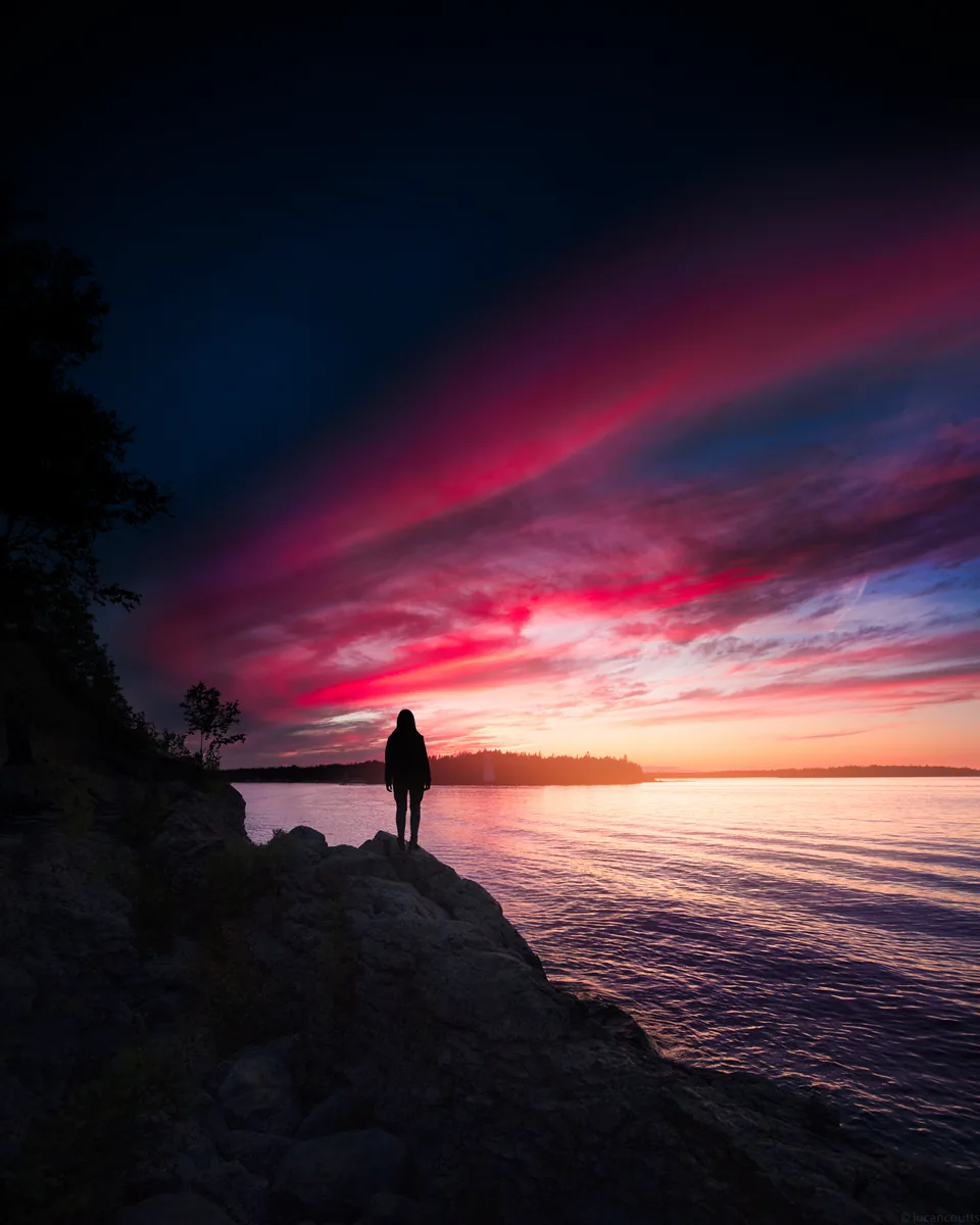 A figure on a rocky shore in Tobermory, Ontario, watching a pink and red sunset over the lake
