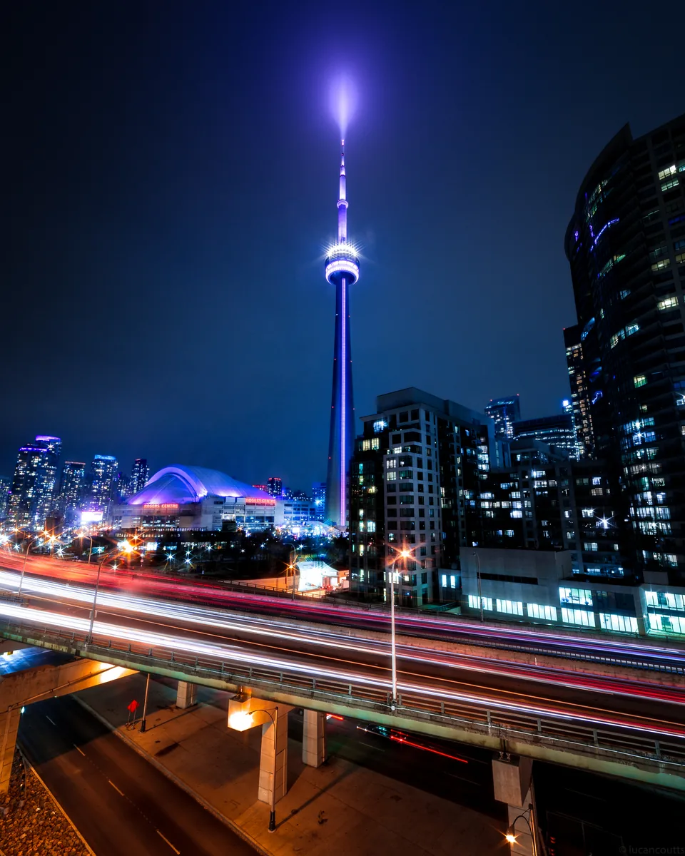 The CN Tower lit purple above car light trails on the Gardiner Expressway at night