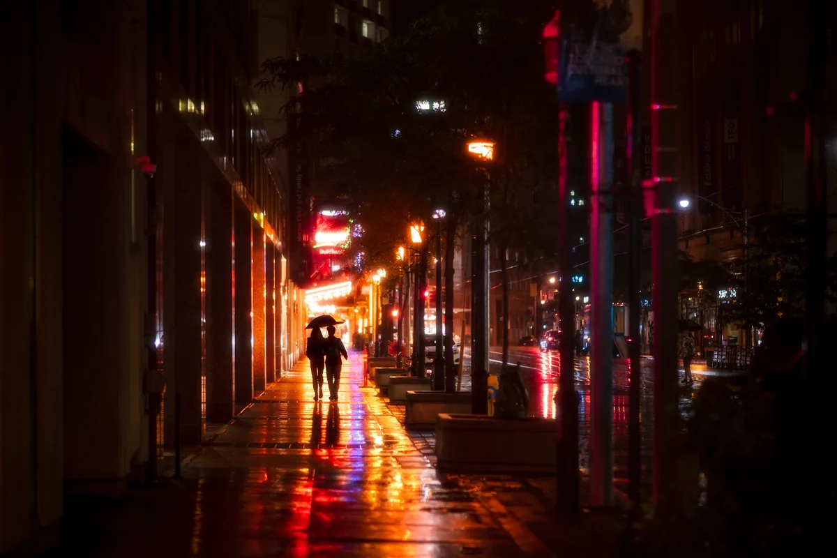 Shelter, a couple sharing an umbrella on a rain-soaked College Street sidewalk in Toronto, neon and streetlights reflected on the pavement