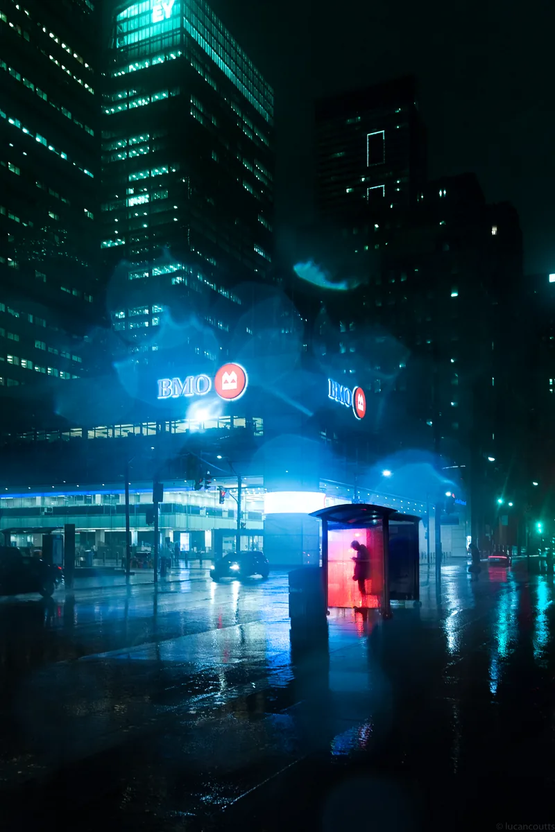 A figure waiting in a glowing bus shelter on a rainy night in Toronto's Financial District