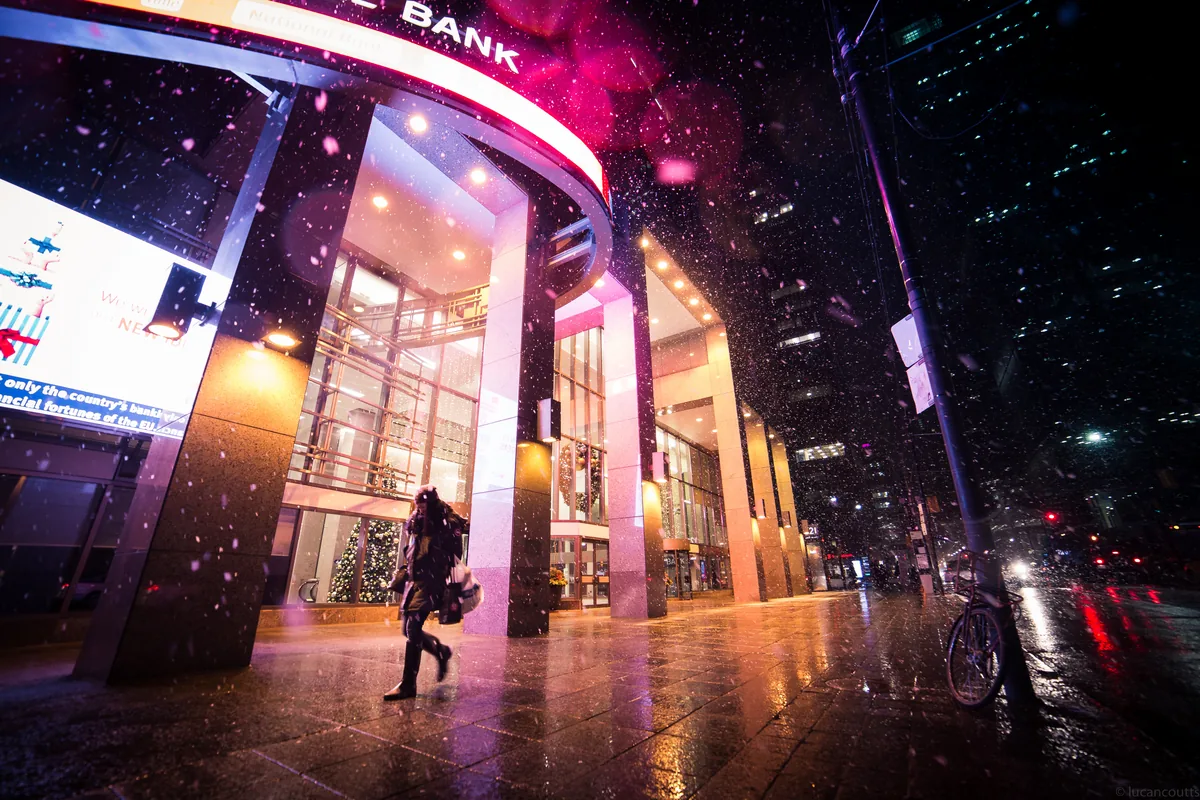 A person walking past a glowing bank entrance on a snowy night in Toronto's Financial District