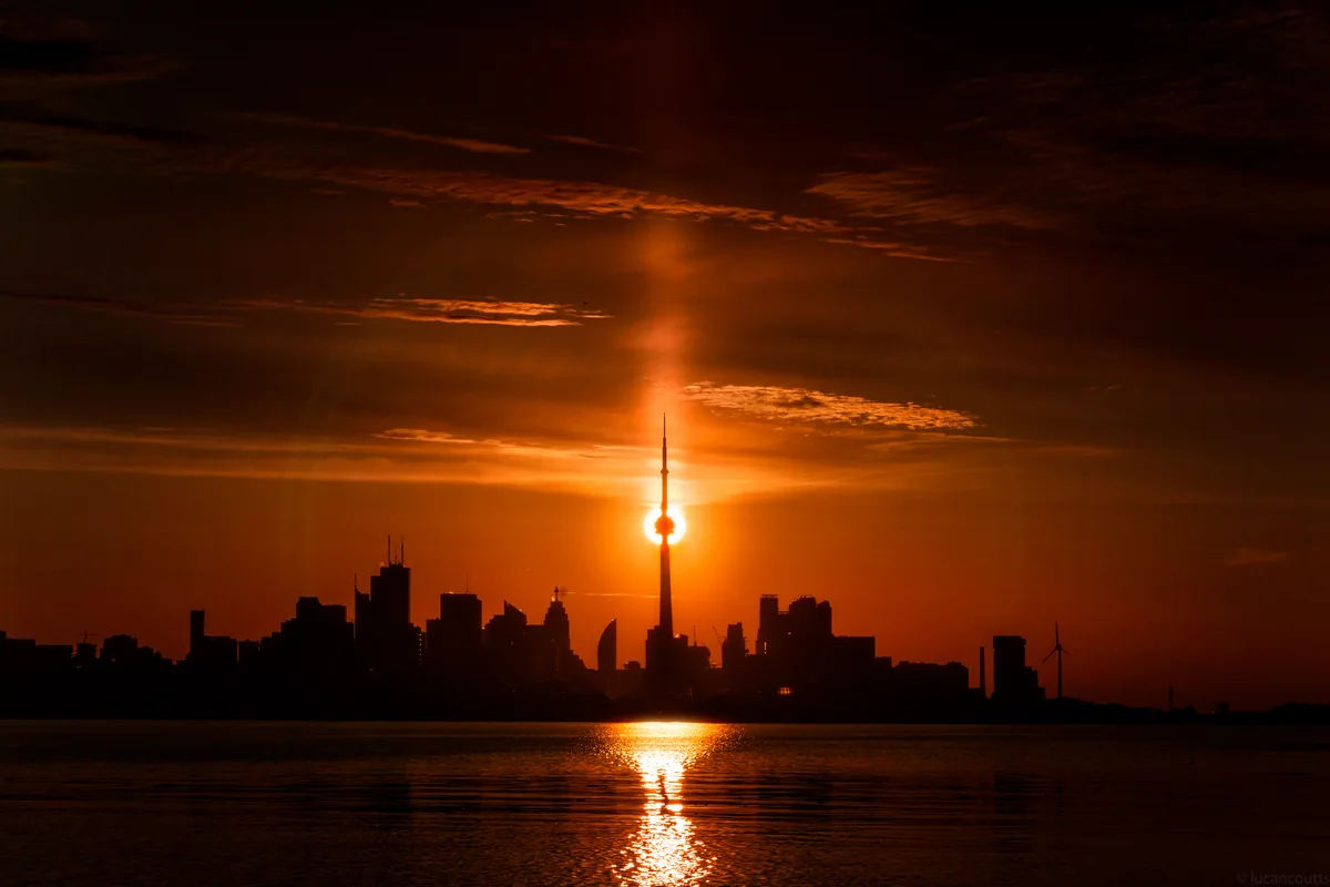 The Toronto skyline in silhouette with the sun setting directly behind the CN Tower, reflected across Lake Ontario